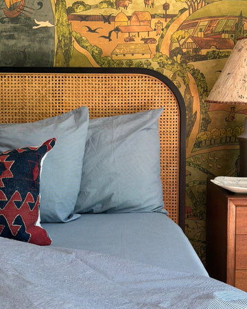 Bedroom with wicker headboard, blue bedding, and decorative pillows against a patterned wall. 100% Organic percale cotton.