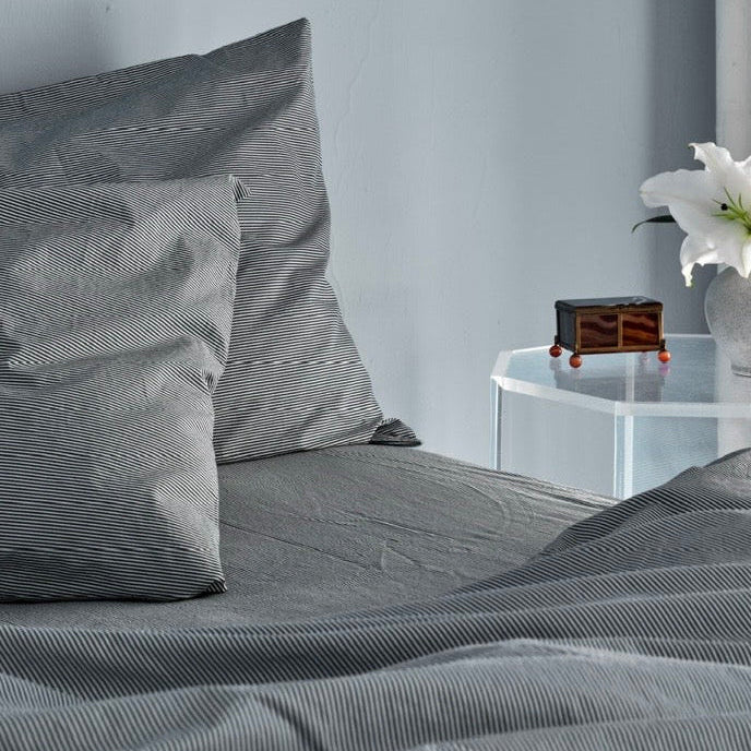 Blue bedding set with striped pillows on a bed, next to a nightstand with a decorative item and flowers. 100% Organic percale cotton.