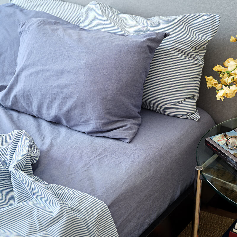 Gray bedding with striped pillows and a side table with a book and flowers.100% Organic percale cotton bed sheets. 