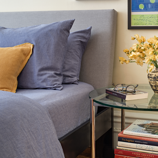 Bedroom with chambray sheets bed, glass nightstand, books, and yellow flowers. 100% Organic cotton bed sheets. 