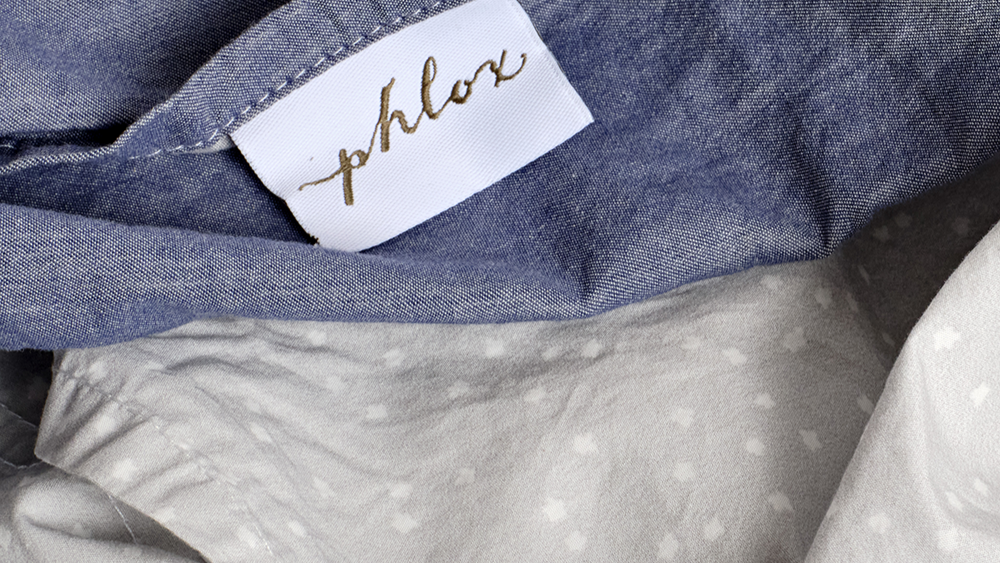 Close-up of a chambray  fabric with a 'phlox' label on a textured background. 100% Organic percale cotton bed sheets. 