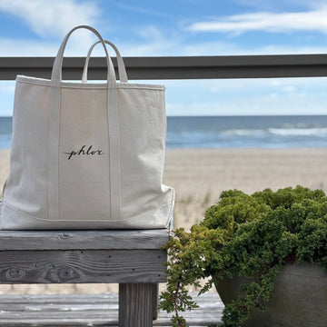 Beige tote bag with 'phlor' branding on a wooden bench by the beach.